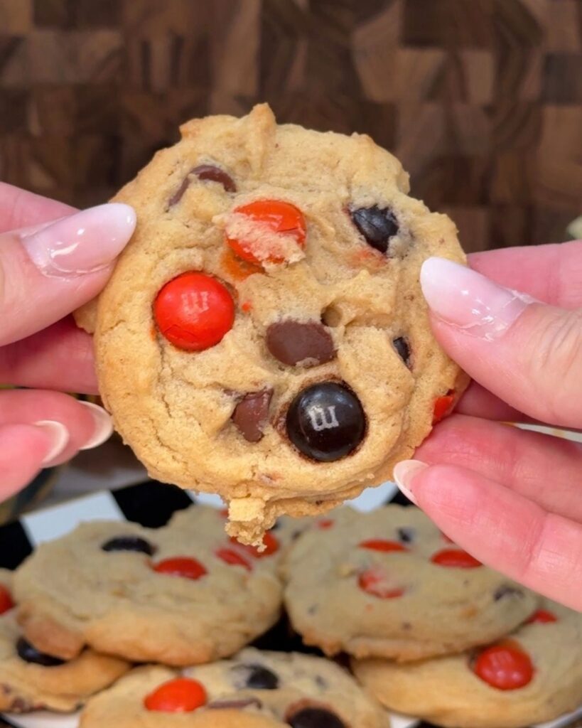 Halloween chocolate chip M&M cookie held between two fingers with chocolate chips and orange candies visible.