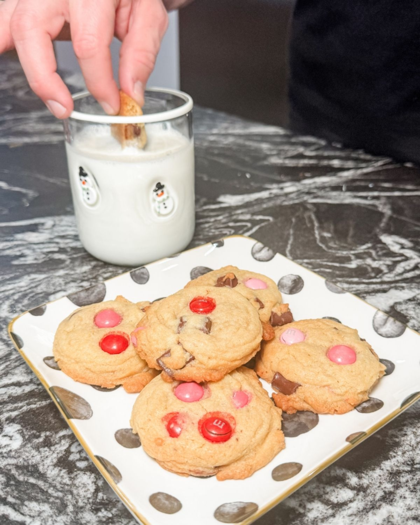 Hand dipping a chocolate chip M&M cookie into a glass of milk beside a plate of cookies.