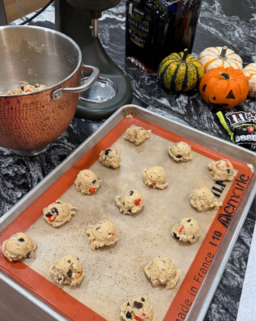 Scoops of chocolate chip M&M cookie dough spaced on a silicone-lined baking sheet.