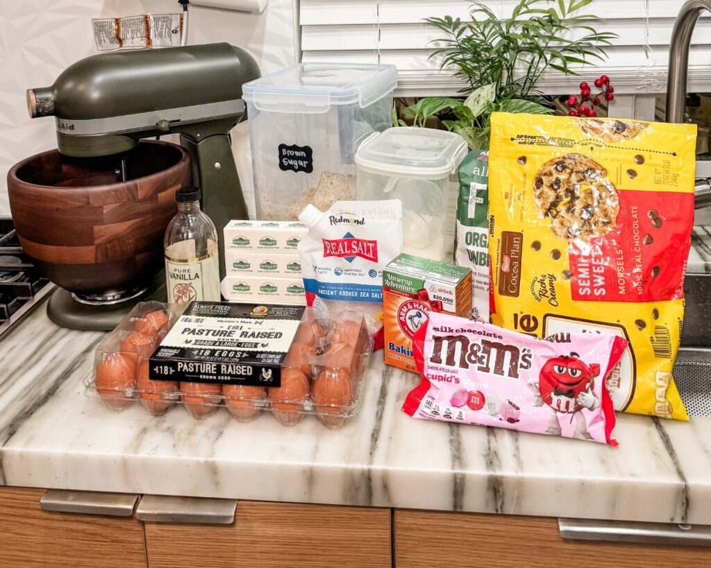 Ingredients for chocolate chip M&M cookies arranged on a kitchen counter beside a stand mixer.