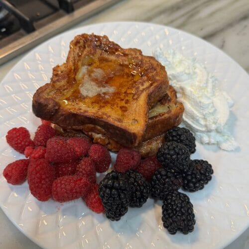 Croissant French toast topped with syrup, raspberries, blackberries, and whipped cream on a white plate