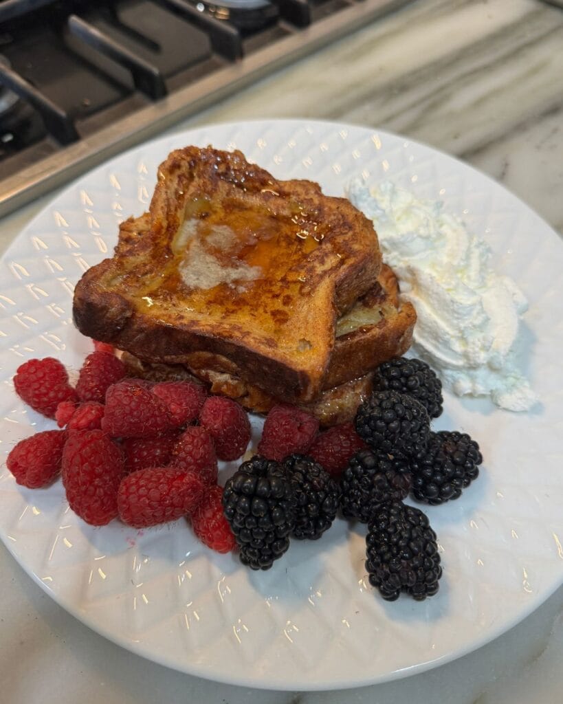 Croissant French toast topped with syrup, raspberries, blackberries, and whipped cream on a white plate