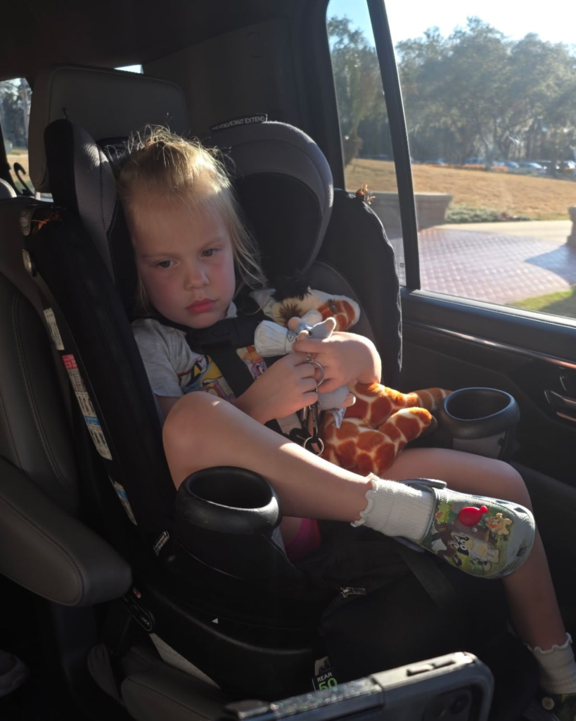 Young child in a car seat holding stuffed animals and travel toys on the way to Disney World.