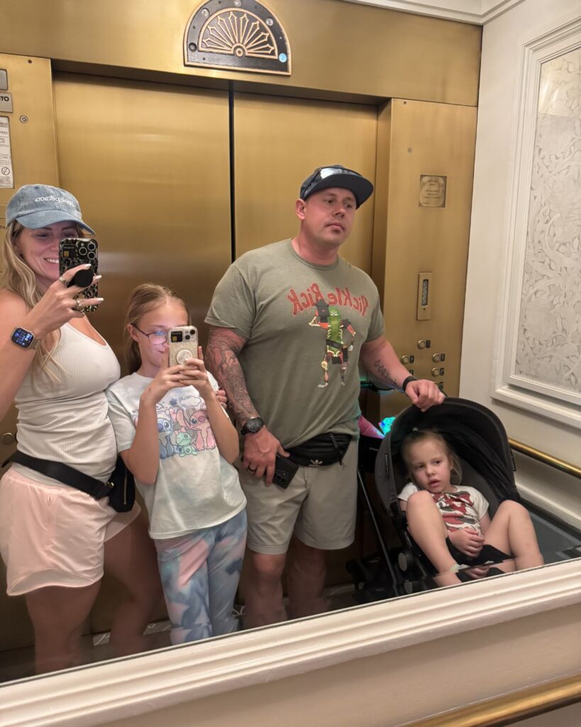 Family mirror selfie in a Disney resort elevator with stroller and park bags.