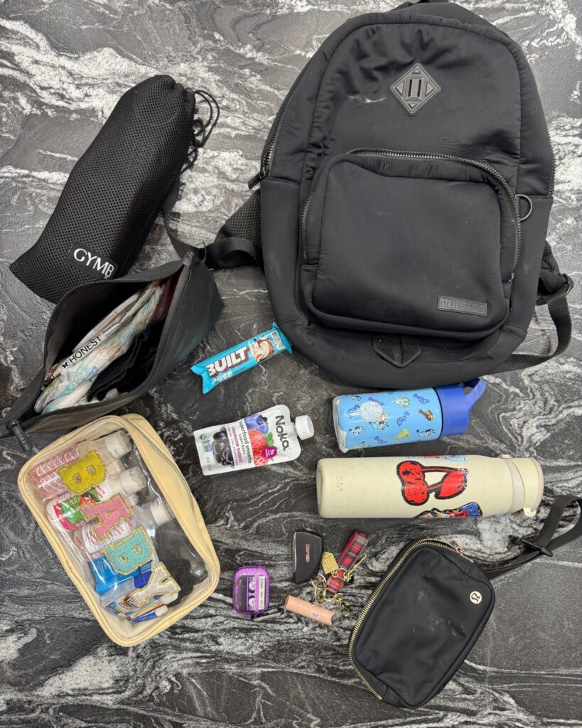 Gym bag essentials for moms including a black backpack, toddler supplies, toiletries, snacks, water bottles, and a belt bag