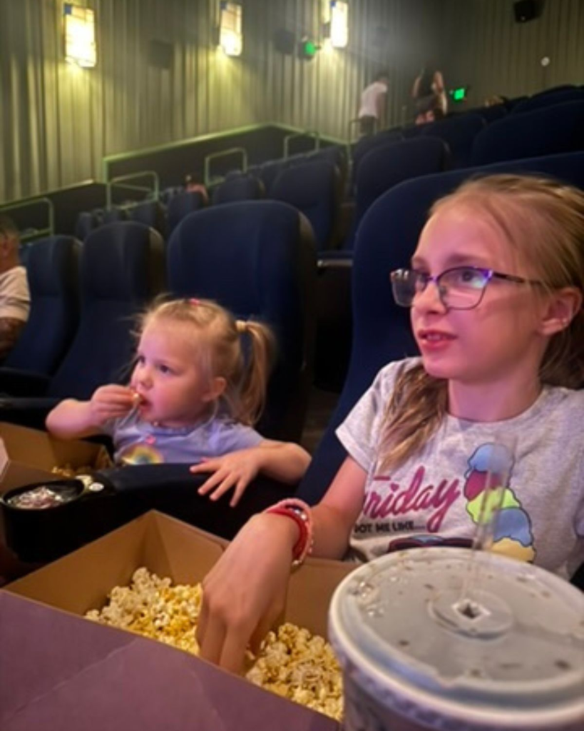 Two sisters sitting in a movie theater eating popcorn during a simple summer outing for kids.