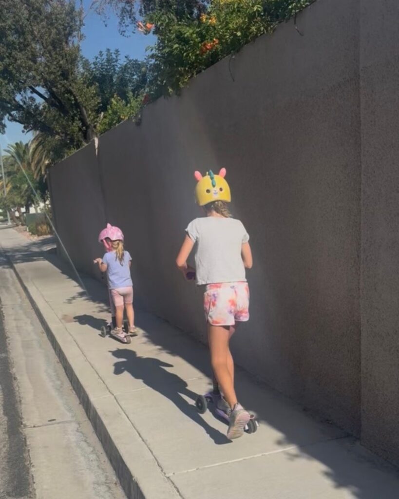 Two girls riding scooters down a neighborhood sidewalk on a sunny summer day.