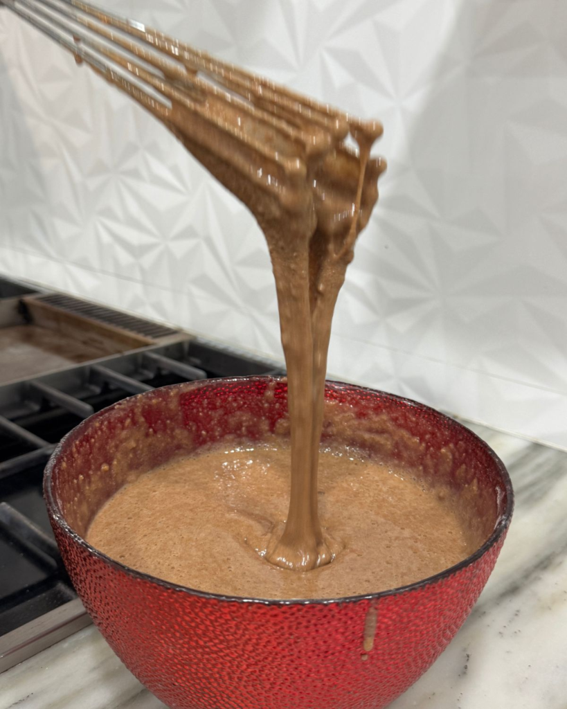 Thick chocolate protein pancake batter falling from a whisk into a mixing bowl.