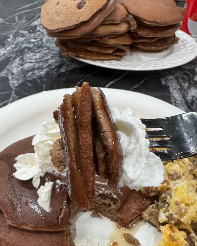 Fork holding a bite of chocolate protein pancake topped with whipped cream and syrup, with a stack of pancakes in the background.