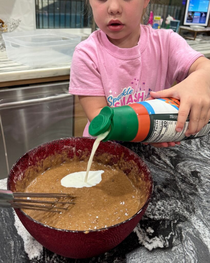 Child pouring coffee creamer into pancake batter while helping prepare protein pancakes.