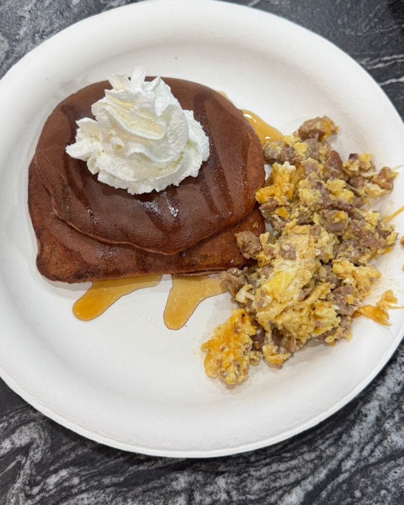 Chocolate protein pancakes served with whipped cream, syrup, scrambled eggs, and sausage.