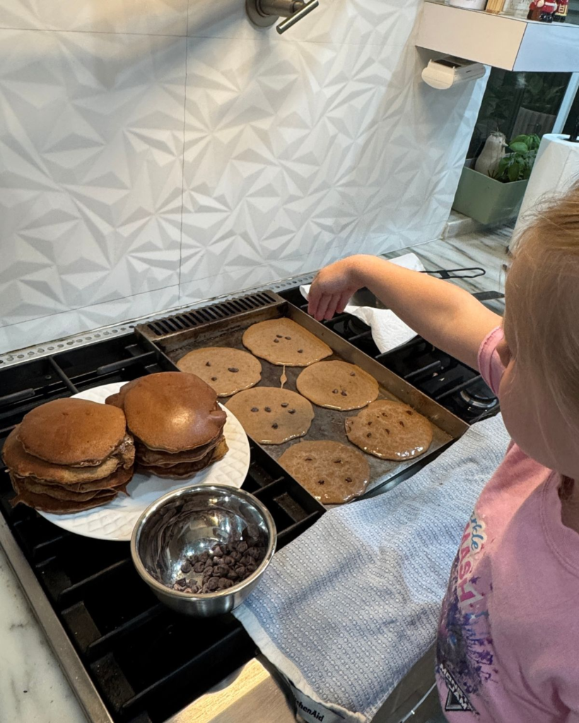 Young girl helping make chocolate protein pancakes with a large stack of finished pancakes nearby.