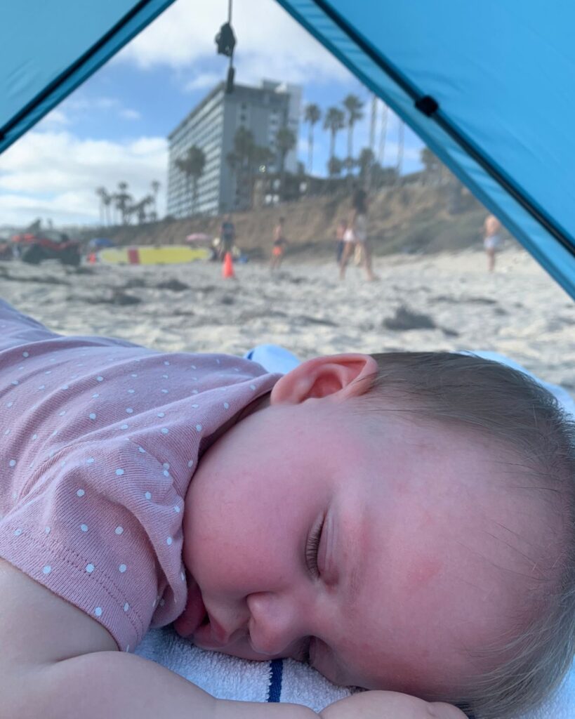 Sleeping toddler inside a beach tent with Pacific Terrace Hotel visible on the bluff behind.