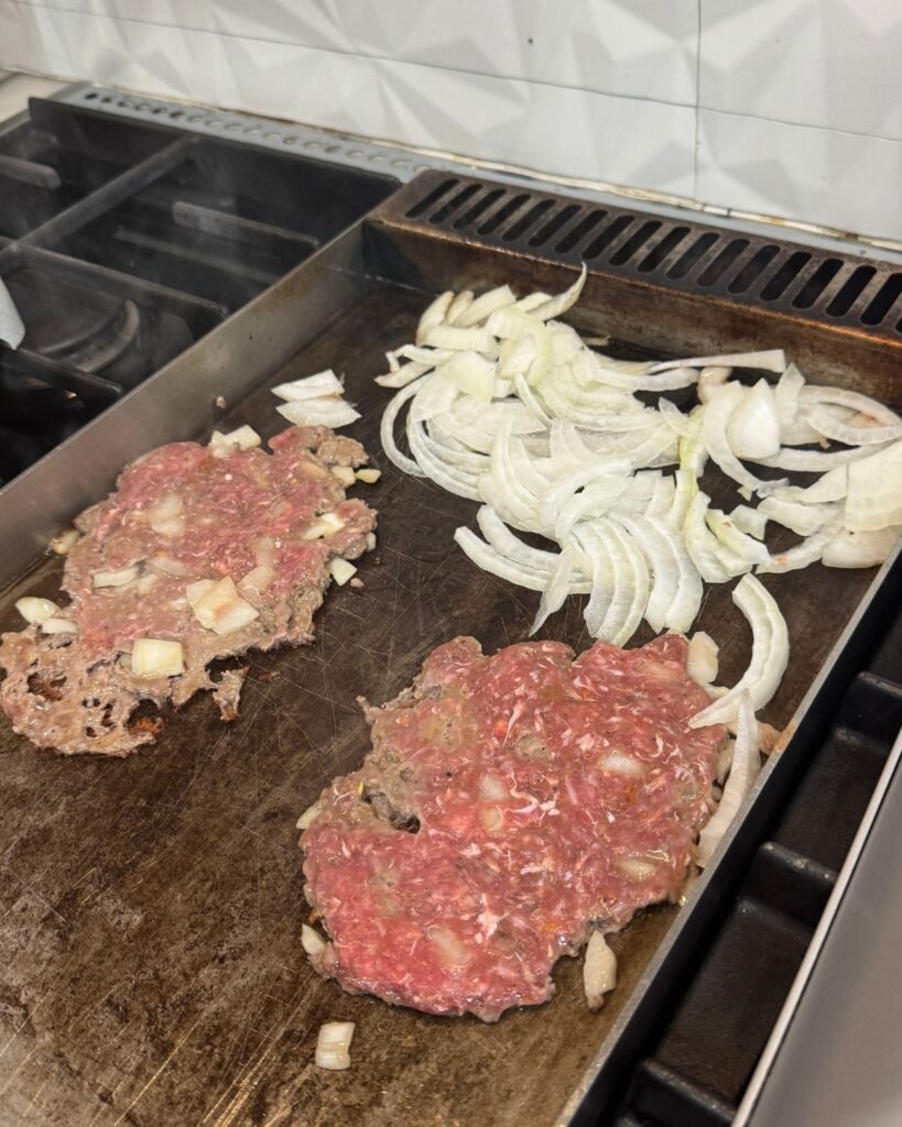 Thin smash burger patties cooking on a flat griddle with sliced onions