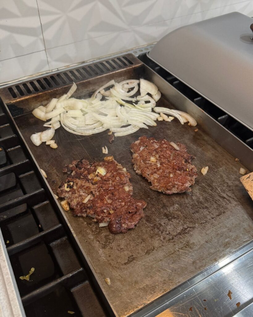 Smash burger patties cooking beside sliced onions on a flat stovetop griddle