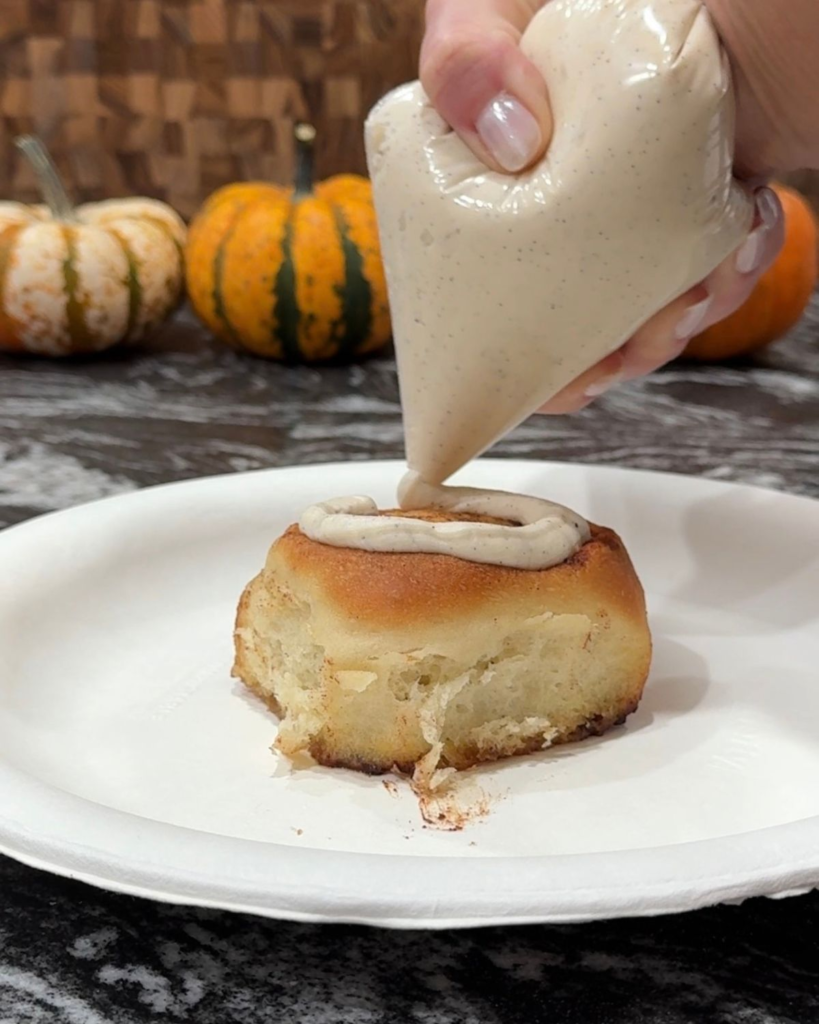 Piping cream cheese frosting onto a freshly baked cinnamon roll
