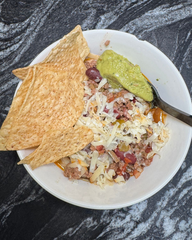 Bowl of slow cooker chili topped with cheese and avocado and served with tortilla chips